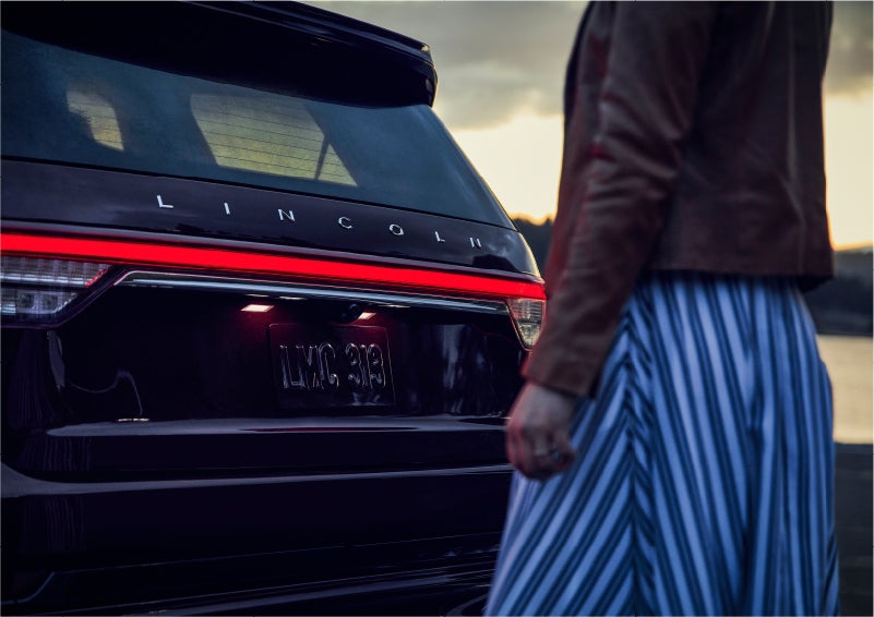 A person is shown near the rear of a 2023 Lincoln Aviator® SUV as the Lincoln Embrace illuminates the rear lights | Lincoln of Cutler Bay in Miami FL